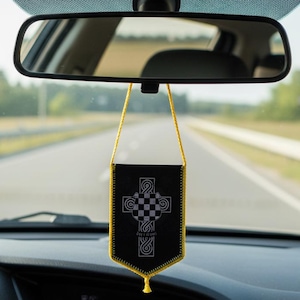 Puede incluir: Un ambientador de coche negro con ribete dorado y un diseño de cruz decorativa. El ambientador cuelga del espejo retrovisor interior de un coche, con una carretera visible en el fondo.