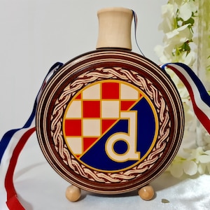 May include: A circular wooden flask with a decorative design. The flask has a red, white, and blue chequered pattern and a blue and white design. It has a wooden stopper and is decorated with red, white, and blue ribbons.