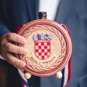 Könnte beinhalten: Eine runde Holzflasche mit dem kroatischen Wappen. Die Flasche hat einen rot-weiß karierten Schild, einen dekorativen Rand und ein rot-weiß-blaues Band. Der Text "BOG I HRVATI" ist sichtbar.