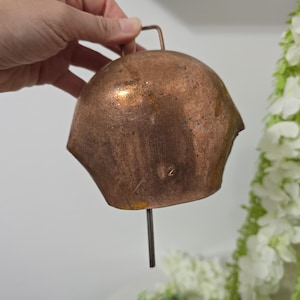 May include: A copper-colored bell with a handle and a clapper. The bell has a weathered appearance with visible wear and tear. The number "12" is stamped on the bell. The bell is held by a hand.