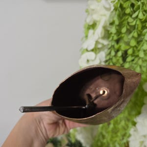 May include: A rustic, bronze-colored bell with a dark interior and a clapper. The bell is held in a hand, with a backdrop of white and green floral arrangements. The bell's shape is reminiscent of a leaf.