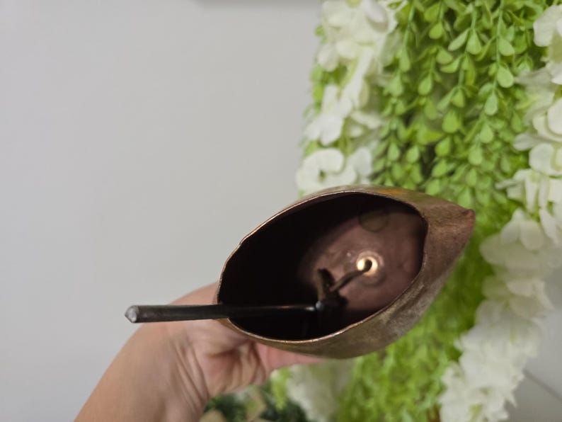 May include: A rustic, bronze-colored bell with a dark interior and a clapper. The bell is held in a hand, with a backdrop of white and green floral arrangements. The bell's shape is reminiscent of a leaf.