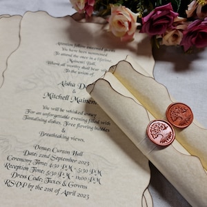 Puede incluir: Un rollo de invitación de boda con un sello de cera con un diseño de árbol. La invitación es para Aisha De y Mitchell Mainem, e incluye la fecha, la hora y el lugar de la ceremonia y la recepción de la boda. El código de vestimenta es esmoquin y vestidos de noche. RSVP antes del 21 de abril de 2023.