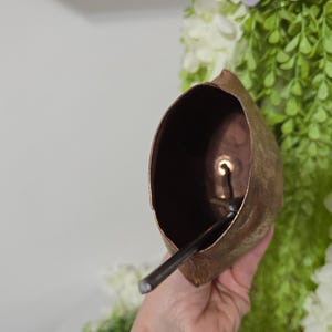 May include: A copper-colored bell with a dark interior and a clapper. The bell is held in a hand, with green and white foliage in the background. The bell has a rustic, aged appearance.