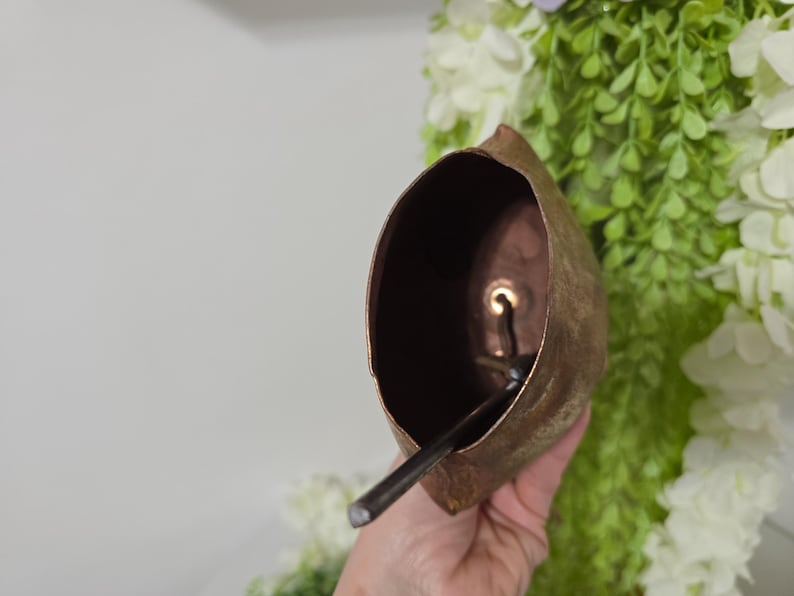 May include: A copper-colored bell with a dark interior and a clapper. The bell is held in a hand, with green and white foliage in the background. The bell has a rustic, aged appearance.