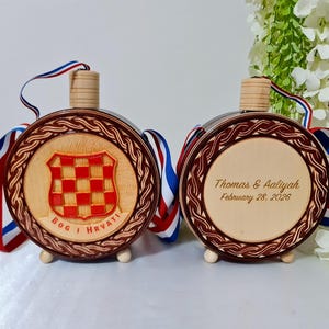 Puede incluir: Dos frascos redondos de madera con centros de madera clara y bordes de madera oscura con patrón trenzado. Un frasco muestra un escudo a cuadros rojos y blancos con el texto BOG I HRVATI. El otro tiene el texto Thomas & Aaliyah February 28, 2026. Ambos tienen cintas rojas, blancas y azules.