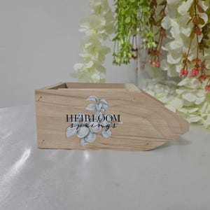 May include: A light-coloured wooden box with a slanted front. The box has the words "HEIRLOOM springs" printed on the side, along with a floral design. The box is sitting on a white surface.