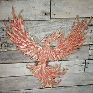 May include: A wooden phoenix wall art piece with outstretched wings. The bird is primarily red with gold accents, and the wings and tail feathers have intricate details. The artwork is mounted on a rustic, weathered wooden background.