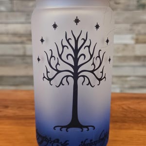 May include: A frosted glass jar with a wooden lid, featuring a black silhouette of the White Tree of Gondor with stars and the inscription "One Ring to rule them all" in Elvish script. The jar has a blue ombre effect at the bottom.