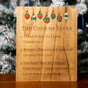 Op de afbeelding: Een houten bord met de tekst "The Code of Elves" en drie regels: Behandel elke dag als Kerstmis, Er is ruimte voor iedereen op de nette lijst, en De beste manier om kerstvreugde te verspreiden is hardop te zingen zodat iedereen het kan horen. Het bord is versierd met zes kleurrijke ornamenten.