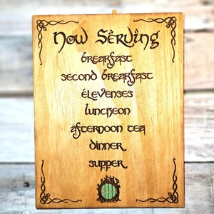 May include: Wooden sign with the words "Now Serving" and meal times, including "Breakfast," "Second Breakfast," "Elevenses," "Luncheon," "Afternoon Tea," "Dinner," and "Supper." The sign has decorative borders and a green door illustration.