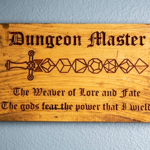 May include: A wooden sign with the text "Dungeon Master" in a decorative font. The sign also features a sword made of dice and the text "The Weaver of Love and Fate. The gods fear the power that I wield."