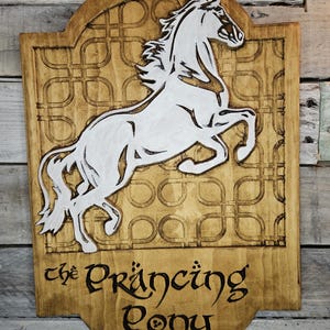 May include: A wooden sign with a white silhouette of a horse rearing up. The sign is painted brown and has a decorative border. The text "the Prancing Pony" is written in black cursive lettering.