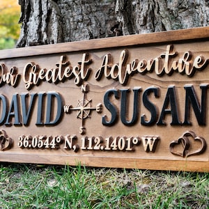 May include: A wooden sign with the text "Our Greatest Adventure" in cursive script. Below the text are the names "DAVID" and "SUSAN" in black lettering, with a compass rose between them. The coordinates "36.0544° N, 112.1401° W" are written below the names. The sign is decorated with two carved hearts.