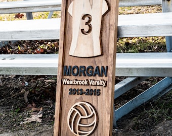 May include: A wooden plaque with a volleyball theme. The plaque features a cutout of a jersey with the number 3, the name "MORGAN", "Westbrook Varsity 2013-2015", and a volleyball. The wood has a dark brown stain.