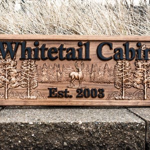 Peut inclure: Une pancarte en bois avec le texte "Whitetail Cabin Est. 2003" grav&eacute; dessus. La pancarte pr&eacute;sente une image grav&eacute;e d'un cerf de Virginie debout dans un d&eacute;cor de for&ecirc;t.