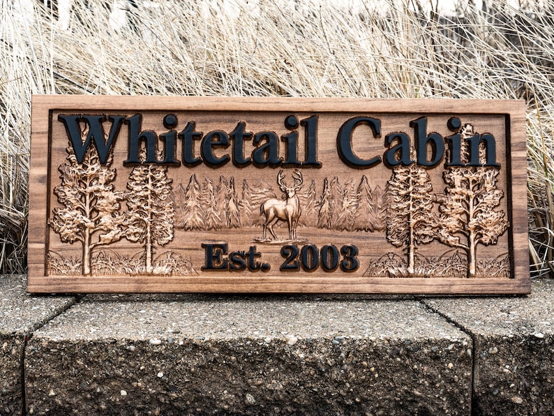 Peut inclure: Une pancarte en bois avec le texte "Whitetail Cabin Est. 2003" grav&eacute; dessus. La pancarte pr&eacute;sente une image grav&eacute;e d'un cerf de Virginie debout dans un d&eacute;cor de for&ecirc;t.