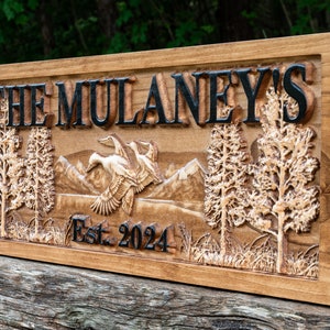 May include: A wooden sign with the text "The Mulaney's Est. 2024" carved into it. The sign features a carved mountain scene with two trees and a flock of geese flying over the mountains.