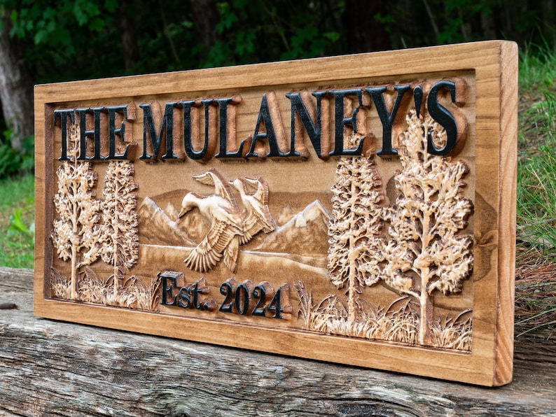 May include: A wooden sign with the text "The Mulaney's Est. 2024" carved into it. The sign features a carved mountain scene with two trees and a flock of geese flying over the mountains.