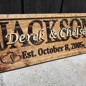 May include: A wooden sign with the name "Jackson" carved in dark brown, with "Derek & Chelsey" in lighter wood. The date "Est. October 8, 2005" is below, with two intertwined hearts. The sign is rectangular and has a dark brown border.