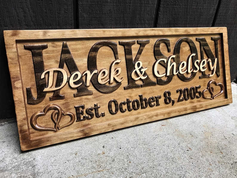 May include: A wooden sign with the name "Jackson" carved in dark brown, with "Derek & Chelsey" in lighter wood. The date "Est. October 8, 2005" is below, with two intertwined hearts. The sign is rectangular and has a dark brown border.
