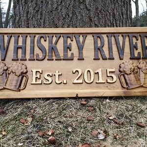 May include: A wooden sign with the text "Whiskey River Est. 2015" and two carved beer mugs on either side.