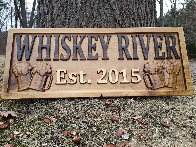 May include: A wooden sign with the text "Whiskey River Est. 2015" and two carved beer mugs on either side.