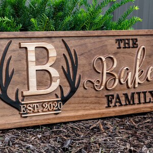May include: A wooden sign with the text "The Baker Family Est. 2020" in black lettering. The sign features a large, carved wooden letter "B" with antlers on either side.