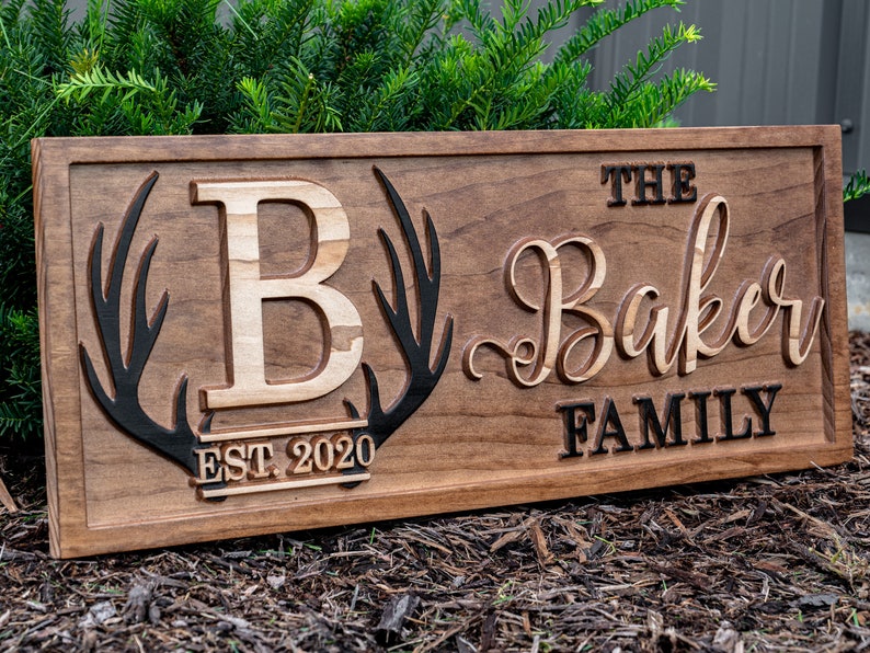 May include: A wooden sign with the text "The Baker Family Est. 2020" in black lettering. The sign features a large, carved wooden letter "B" with antlers on either side.