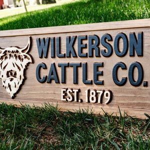 May include: A wooden sign with the text "Wilkerson Cattle Co. Est. 1879" in black raised letters. A carved wooden image of a Highland cow is on the left side of the sign.