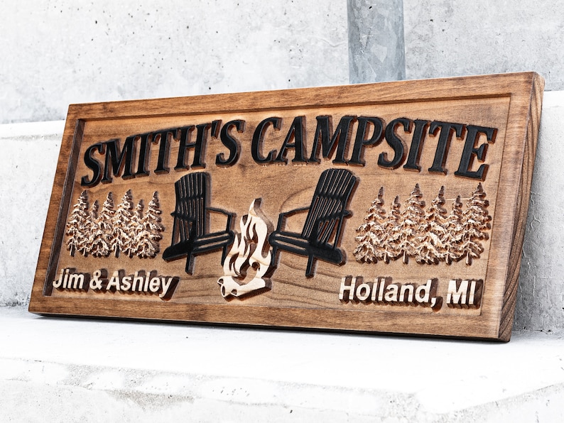 Puede incluir: Un letrero de madera con el texto "SMITH'S CAMPSITE" en letras negras. El letrero tambi&eacute;n incluye los nombres "Jim & Ashley" y "Holland, MI" en letras negras. El letrero presenta una imagen tallada de dos sillas Adirondack enfrentadas con una fogata en el medio.