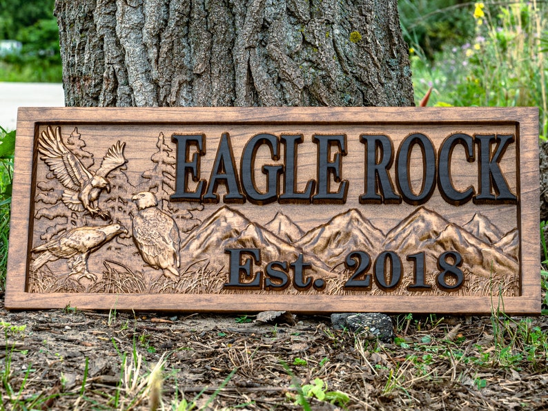 May include: A wooden sign with a carved design of eagles and mountains. The sign reads "Eagle Rock Est. 2018".