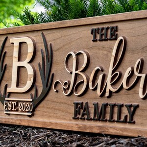 May include: A wooden sign with the text "The Baker Family Est. 2020" in a rustic font. The sign features a large "B" surrounded by antlers.