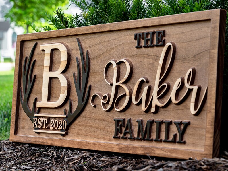 May include: A wooden sign with the text "The Baker Family Est. 2020" in a rustic font. The sign features a large "B" surrounded by antlers.