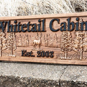 Peut inclure: Une pancarte en bois avec les mots "Whitetail Cabin" grav&eacute;s en lettres noires. La pancarte pr&eacute;sente &eacute;galement une image grav&eacute;e d'un cerf de Virginie dans un d&eacute;cor forestier et le texte "Est. 2003".