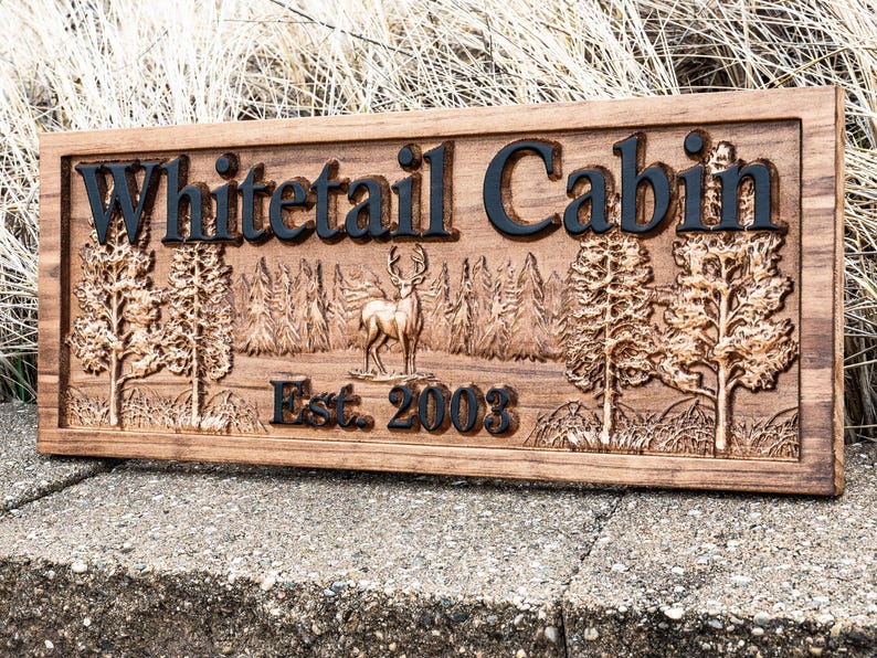 Peut inclure: Une pancarte en bois avec les mots "Whitetail Cabin" grav&eacute;s en lettres noires. La pancarte pr&eacute;sente &eacute;galement une image grav&eacute;e d'un cerf de Virginie dans un d&eacute;cor forestier et le texte "Est. 2003".