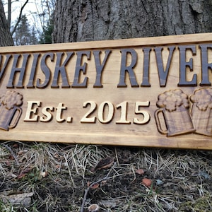 May include: Wooden sign with the text "Whiskey River Est. 2015" and two carved beer mugs.