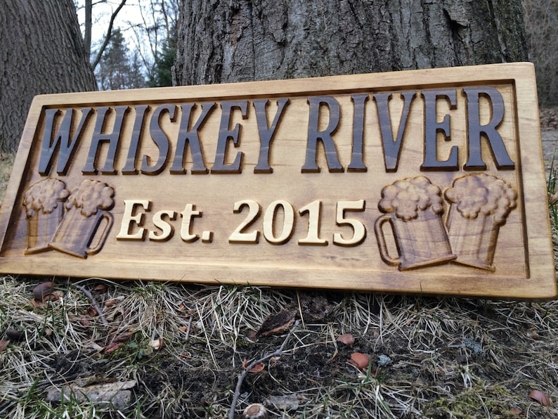 May include: Wooden sign with the text "Whiskey River Est. 2015" and two carved beer mugs.