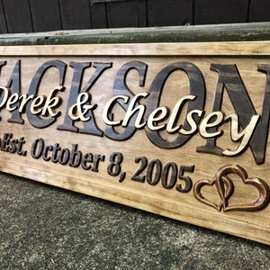 May include: A wooden sign with the name "Jackson" in dark brown, and "Derek & Chelsey" in lighter wood. The sign includes "Est. October 8, 2005" and two heart designs. The sign is framed with a light brown border.