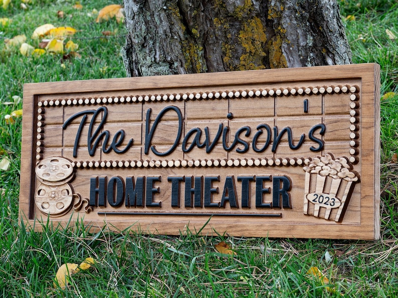 Pu&ograve; includere: Un cartello in legno con il testo "The Davisons' Home Theater 2023" in lettere nere. Il cartello &egrave; decorato con una bobina di pellicola e un secchiello di popcorn.