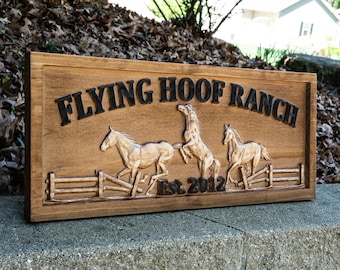 Può includere: Targa in legno marrone con la scritta nera "FLYING HOOF RANCH", raffigurante tre cavalli e una staccionata. È presente anche il testo "Est. 2012". La cornice è scura.