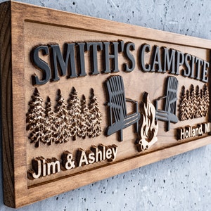 Puede incluir: Un letrero de madera con el texto "Smith's Campsite" en letras negras. El letrero tambi&eacute;n presenta una imagen tallada de dos sillas, una fogata y una hilera de pinos. Los textos "Jim & Ashley" y "Holland, MI" tambi&eacute;n est&aacute;n tallados en el letrero.