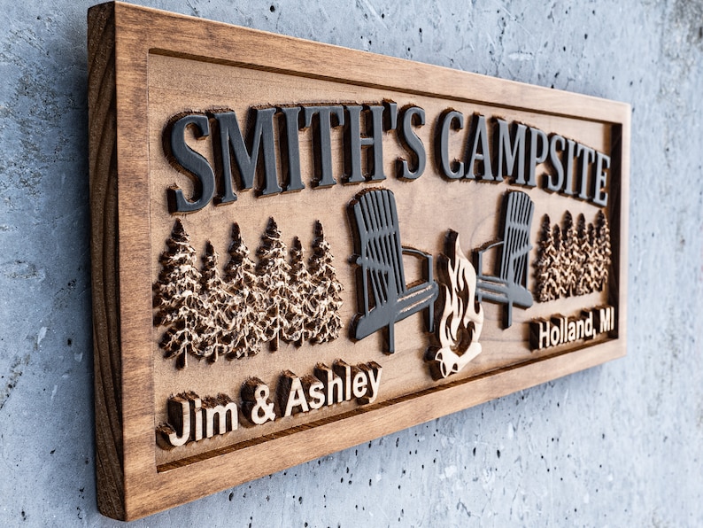 Puede incluir: Un letrero de madera con el texto "Smith's Campsite" en letras negras. El letrero tambi&eacute;n presenta una imagen tallada de dos sillas, una fogata y una hilera de pinos. Los textos "Jim & Ashley" y "Holland, MI" tambi&eacute;n est&aacute;n tallados en el letrero.