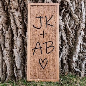 May include: A rectangular wooden sign with a textured, brown surface. The sign features the initials "JK" above "+" and "AB", with a heart symbol below. The sign is framed in a light brown wood.