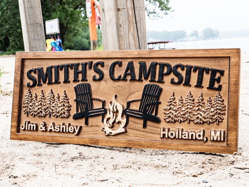 Puede incluir: Un letrero de madera con el texto "SMITH'S CAMPSITE" en letras negras. El letrero tambi&eacute;n presenta dos siluetas negras de sillas Adirondack enfrentadas con una fogata entre ellas. El texto "Jim & Ashley" est&aacute; en el lado izquierdo del letrero y "Holland, MI" est&aacute; en el lado derecho.