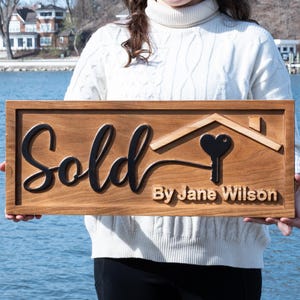 May include: A wooden sign with the word "Sold" in black script. The sign features a house outline with a heart-shaped key and the text "By Jane Wilson". The sign is brown.