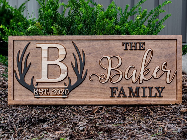 May include: A wooden sign with the text "The Baker Family" in a cursive font. The sign also features a large letter "B" and a pair of antlers. The text "Est. 2020" is below the letter "B".