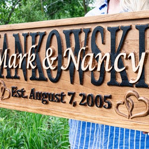 May include: A wooden sign featuring the names "Mark & Nancy" in a light colour, set against a dark background, with the surname "Jankowski" above. The sign also includes the date "Est. August 7, 2005" and heart-shaped carvings.