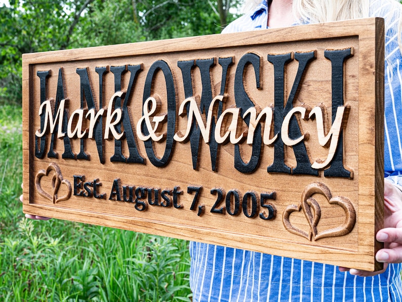 May include: A wooden sign featuring the names "Mark & Nancy" in a light colour, set against a dark background, with the surname "Jankowski" above. The sign also includes the date "Est. August 7, 2005" and heart-shaped carvings.