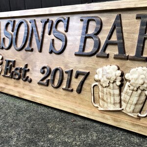May include: A wooden sign with the text "Jason's Bar Est. 2017" in black lettering. The sign features two carved wooden beer mugs with foam on top.